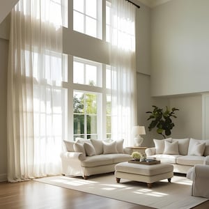 May include: A living room featuring two cream-coloured sofas and a matching ottoman arranged on a light-coloured rug. Tall, sheer white curtains hang from the ceiling, covering large multi-pane windows that allow natural light to fill the space.