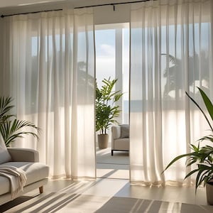May include: Cream-coloured, sheer curtains hang from a dark rod, framing a bright outdoor view. Sunlight filters through the fabric, illuminating the room. Potted plants and a neutral sofa contribute to the airy, natural aesthetic.