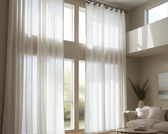 May include: Long, sheer white curtains hang from a dark rod, draping in front of large windows. The curtains create a soft, airy feel. The room has a neutral colour palette with a sofa, coffee table, and a vase of greenery.