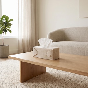 Peut inclure: Une boîte à mouchoirs rectangulaire en feutre beige avec un design plié fixé par un petit bouton métallique sur le côté. La housse est fabriquée dans un matériau doux et texturé et contient une pile de mouchoirs en papier blancs sur le dessus.