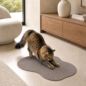 May include: A brown tabby cat stretches its body while standing on an irregular, cloud-shaped grey felt mat. The mat features a smooth, flat surface with a small, circular metallic accent near the edge, providing a soft area for the cat to rest.