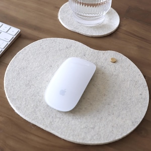Peut inclure: Un tapis de bureau en feutre gris clair de forme ovale irrégulière, doté d'un petit accent métallique de couleur laiton sur le côté droit. Une souris d'ordinateur blanche est posée sur le tapis, composé d'une matière texturée et douce.