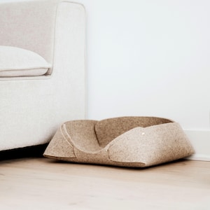 Puede incluir: Una cama para mascotas de fieltro beige con un diseño abierto y de perfil bajo. La estructura está formada por paneles superpuestos de fieltro prensado, asegurados con un pequeño cierre metálico en el lateral, creando una forma redondeada.