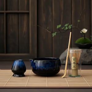 May include: A Japanese tea ceremony set featuring a blue and black ceramic bowl, a smaller matching cup, a bamboo whisk, and a bamboo scoop. The set is arranged on a bamboo mat against a dark wooden backdrop.