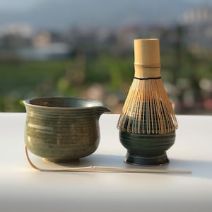 Könnte beinhalten: Ein Matcha-Teeset mit einer dunkelgrünen Keramikschale, einem Bambusbesen und einem Löffel. Der obere Teil des Schneebesens ist hellbraun. Das Set steht auf einer weißen Oberfläche, ideal für Matcha-Tee.