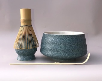 May include: A matcha tea set featuring a speckled teal ceramic bowl and whisk base, a bamboo whisk, and a bamboo scoop. The bowl has a white interior. The whisk is made of bamboo and has a natural wood finish.