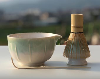 May include: A matcha tea set featuring a light green and white ceramic bowl with a spout, a bamboo whisk, and a bamboo scoop. The bowl has vertical grooves and a curved handle. The whisk is light brown and the scoop is a light beige color.