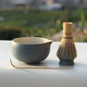 May include: A matcha tea set featuring a speckled blue ceramic bowl with a spout, a bamboo whisk, and a bamboo scoop. The bowl has a white interior. The whisk has a dark blue base. The set is designed for preparing matcha tea.