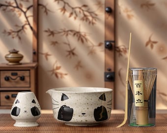 May include: A matcha tea set featuring a ceramic bowl, whisk, and a small holder, all decorated with a cat face design. The set includes a bamboo whisk and a scoop, perfect for preparing matcha tea. The bowl is speckled with a light beige color.