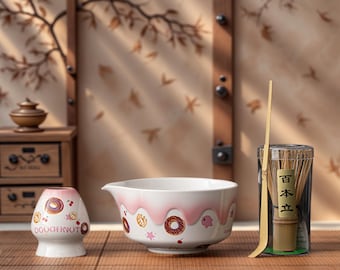 Könnte beinhalten: Ein Keramik-Matcha-Schüssel-Set mit einem rosa und weißen Donut-Design. Das Set enthält eine Schüssel, einen Schneebesen und einen Halter. Auf dem Halter steht das Wort "DOUGHNUT". Der Schneebesen ist aus Bambus.