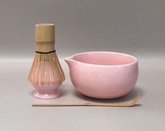 Può includere: Una ciotola per matcha in ceramica rosa con beccuccio, un frullino in bambù e un cucchiaio in bambù. Il frullino è rosa chiaro con un manico in bambù naturale. La ciotola e il frullino sono su una superficie grigia.