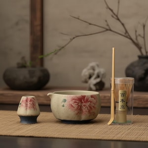 May include: A ceramic matcha tea set featuring a bowl with a floral design, a whisk, and a small tea cup. The set includes a bamboo whisk and a clear container. The tea set is on a bamboo mat.