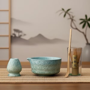 May include: A matcha tea set with a light blue ceramic bowl, a whisk, a whisk holder, and a bamboo scoop. The set is arranged on a bamboo mat, with a clear container holding the whisk. The background features a serene landscape painting.