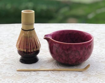 May include: A matcha tea set featuring a burgundy ceramic bowl with a spout, a bamboo whisk, and a bamboo scoop. The whisk has a dark base and light-colored handle. The bowl and whisk are on a white lace tablecloth.