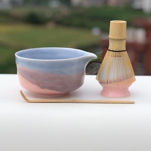Puede incluir: Un juego de té matcha con un cuenco de cerámica de colores pastel con un pico, un batidor de bambú y una cuchara de bambú. El cuenco tiene un degradado azul claro, morado y rosa. El batidor y la base son rosas.