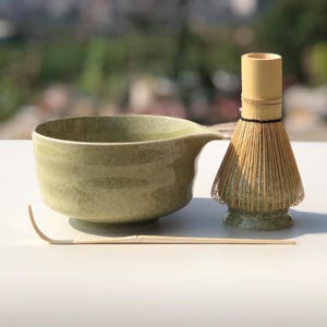 May include: A matcha tea set featuring a green ceramic bowl with a spout, a bamboo whisk, and a bamboo scoop. The bowl has a textured surface and a light green color. The whisk is made of thin bamboo tines.