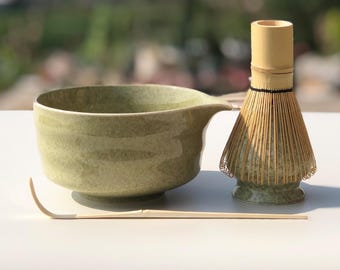 May include: A matcha tea set featuring a green ceramic bowl with a spout, a bamboo whisk, and a bamboo scoop. The bowl has a textured surface and a light green color. The whisk is made of thin bamboo tines.