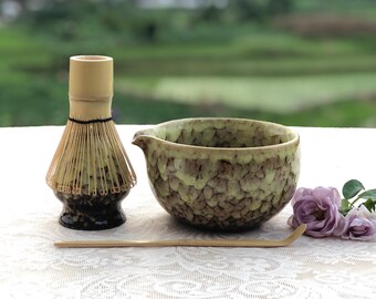 May include: A matcha tea set featuring a bamboo whisk, a ceramic bowl with a spout, and a bamboo scoop. The bowl has a textured, mottled green and brown glaze. The whisk is light brown with a black base. A few purple flowers are in the background.
