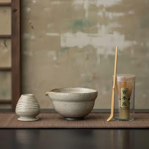 May include: A ceramic matcha tea set with a bowl, whisk, and scoop. The bowl is light beige with a spout. The whisk is in a clear container with Japanese text. The set includes a small, striped ceramic holder.