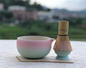 Può includere: Un set da tè matcha con una ciotola in ceramica sfumata rosa e verde, un frullino in bambù e un cucchiaio in bambù. Il frullino ha una base verde chiaro e un manico marrone chiaro. Il set è su una tovaglia di pizzo bianco.