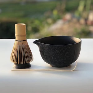 May include: A matcha tea set featuring a bamboo whisk, a black ceramic bowl with a spout, and a bamboo scoop. The whisk has a dark base and light-colored bristles. The bowl has a speckled texture.