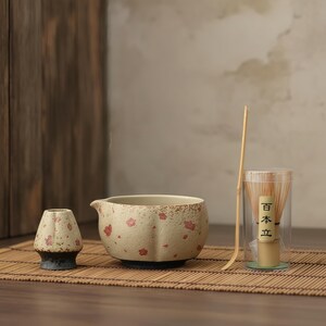 May include: A matcha tea set comprising a beige ceramic bowl with red floral details, a small matching tea cup, a bamboo whisk, and a bamboo scoop. The whisk is in a clear container. The set is on a bamboo mat.