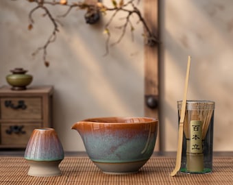 May include: A matcha tea set with a ceramic bowl, whisk, and scoop. The bowl has a brown and teal glaze. The whisk is in a clear container with Japanese text. The set is on a bamboo mat.