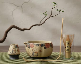 Op de afbeelding: Een Japanse theeset met een beige keramische kom versierd met katten en bloemen, een kleine bijpassende container, een bamboe klopper en een lepel. De set bevat een bamboe klopper in een doorzichtige container.