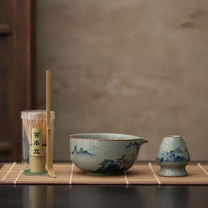 Może przedstawiać: Zestaw do herbaty matcha składający się z ceramicznej miski z dzióbkiem, uchwytu na trzepaczkę i bambusowej trzepaczki z łyżką. Miska i uchwyt na trzepaczkę mają niebieski wzór krajobrazu. Trzepaczka znajduje się w przezroczystym pojemniku z japońskim tekstem.