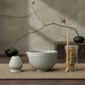 May include: A matcha tea set featuring a light green ceramic bowl with a spout, a small ceramic tea cup, a bamboo whisk, and a bamboo scoop. The whisk and scoop are in a clear plastic container. The set is arranged on a woven mat.