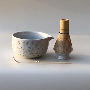 May include: A matcha tea set comprising a speckled ceramic bowl with a spout, a bamboo whisk, and a bamboo scoop. The bowl and whisk base have a speckled design. The whisk is made of bamboo. The set is designed for preparing matcha tea.