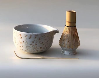 May include: A matcha tea set featuring a speckled ceramic bowl with a spout, a bamboo whisk, and a bamboo scoop. The bowl and whisk base have a speckled design. The whisk is made of bamboo. The set is designed for preparing matcha tea.