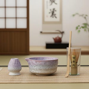 May include: A Japanese tea ceremony set featuring a lavender and white ceramic bowl, a whisk, and a bamboo scoop. The bowl has a textured surface and a spout. The whisk is in a clear container. The set is on a bamboo mat.