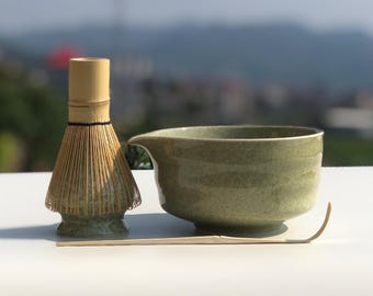 May include: A matcha tea set featuring a bamboo whisk, a ceramic bowl with a spout, and a bamboo scoop. The bowl and whisk base are a speckled green, and the whisk handle is a light brown. The set is arranged on a white surface.