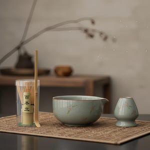 Może przedstawiać: Zestaw do herbaty matcha z jasnoniebieską ceramiczną miską z dzióbkiem, pasującym małym wazonem i bambusową trzepaczką w przezroczystym pojemniku. Zestaw zawiera bambusową łyżkę i jest ułożony na tkanej macie.