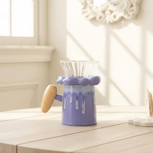 May include: A lilac ceramic mug with a wooden handle and a clear glass top. The mug features a distinctive design with white drips and a wavy rim. The mug is on a light-coloured wooden surface.