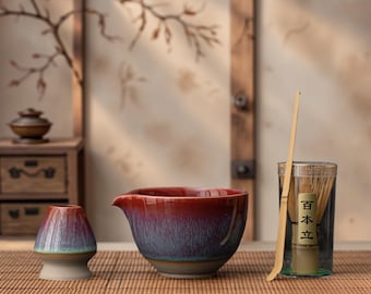 May include: A matcha tea set featuring a red and blue ceramic bowl with a spout, a bamboo whisk, and a ceramic whisk holder. The set is displayed on a bamboo mat, with a glass container holding the whisk.