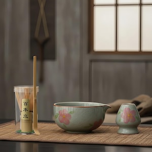 Könnte beinhalten: Ein Matcha-Teeset mit einer Keramikschale mit Ausguss, einem kleinen Keramikständer sowie einem Bambusbesen und -löffel. Die Schale und der Ständer sind hellgrün mit rosa Blumendesigns. Der Schneebesen und der Löffel sind aus Bambus.