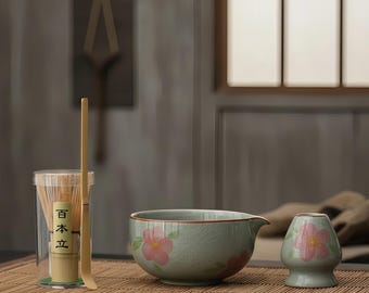 Op de afbeelding: Een matcha-theeset met een keramische kom met een tuit, een kleine keramische standaard en een bamboe klopper en lepel. De kom en standaard zijn lichtgroen met roze bloemmotieven. De klopper en lepel zijn van bamboe.