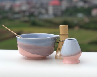 Può includere: Un set da tè matcha in ceramica con schema di colori azzurro, viola e rosa. Il set include una ciotola con manico, un frullino, una tazza piccola e un cucchiaio di bambù. Lo sfondo è sfocato, con un paesaggio verde e degli edifici.