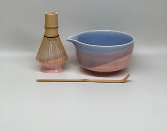 Può includere: Un set da tè matcha con una frusta di bambù, una ciotola in ceramica con sfumatura blu e rosa e un cucchiaio di bambù. La frusta ha una base rosa e un manico marrone chiaro. La ciotola ha un beccuccio.