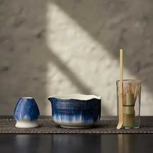 Puede incluir: Un juego de té matcha con un chawan de cerámica azul y blanco, un cuenco de cerámica azul y blanco y un recipiente transparente con un batidor y una cuchara de bambú. El juego está dispuesto sobre una esterilla marrón oscuro.