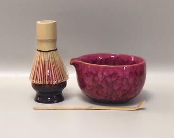 May include: A matcha tea set featuring a bamboo whisk, a deep pink ceramic bowl with a spout, and a bamboo scoop. The whisk has a dark base and light-colored bristles. The bowl has a textured surface.