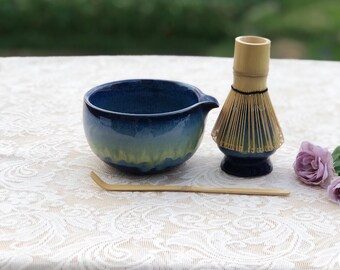 May include: A blue and green ceramic matcha bowl with a spout, a bamboo whisk, and a bamboo scoop. The bowl and whisk sit on a white lace tablecloth. A few purple roses are in the background.