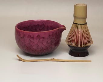 May include: A matcha tea set featuring a textured, magenta-colored ceramic bowl with a spout, a bamboo whisk, and a bamboo scoop. The whisk has a dark brown base and a light brown handle.