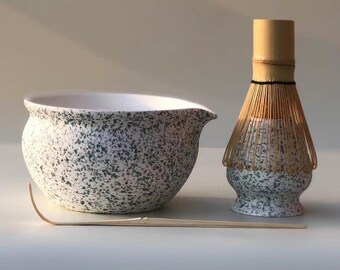 May include: A matcha tea set featuring a speckled ceramic bowl with a spout, a bamboo whisk, and a bamboo scoop. The bowl and whisk base have a white and green speckled pattern. The whisk is made of bamboo.