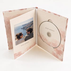 Puede incluir: Una caja de CD abierta con un patrón de acuarela rosa pálido. El panel izquierdo presenta una fotografía de dos personas mirando al océano con el texto OUR SONGS debajo. El panel derecho contiene un CD con el texto OUR SONGS impreso.