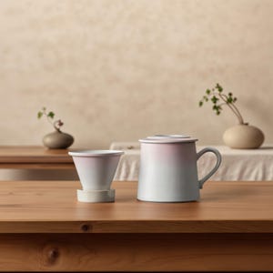 May include: A ceramic coffee dripper and mug set with a pink and grey gradient finish. The dripper sits on a small, round base. The mug has a handle and a lid. The set is on a wooden table.