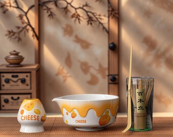 Könnte beinhalten: Ein weißes Keramik-Teeset zum Thema Käse mit einer Schüssel, einer kleinen Tasse und einem Matcha-Besen. Die Schüssel und die Tasse haben gelbe Käse-Akzente und das Wort "CHEESE". Ein Bambusbesen und ein Löffel sind ebenfalls enthalten.