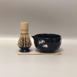 Könnte beinhalten: Ein Matcha-Teeset mit einer schwarzen Keramikschale mit weißen Blumenmotiven, einem Bambusbesen und einem Bambuslöffel. Der Schneebesen ist hellbraun mit schwarzem Sockel. Das Set ist für die Zubereitung von Matcha-Tee.